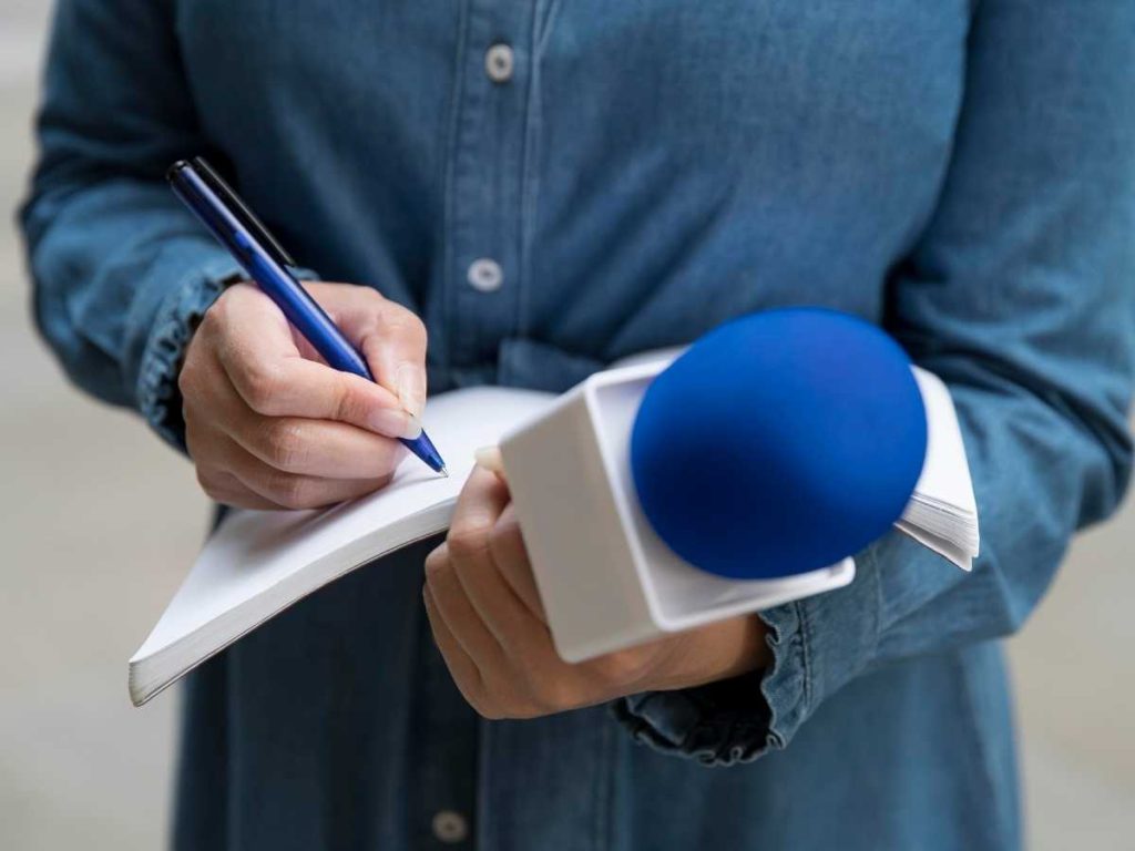 Periodista escribiendo en una libreta con un micrófono azul en la otra mano