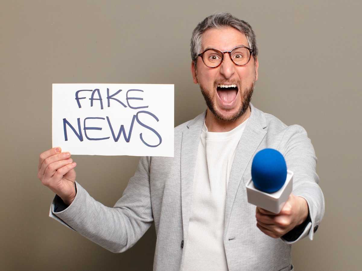 Hombre con gafas y micrófono azul sosteniendo un cartel que dice 'Fake News'