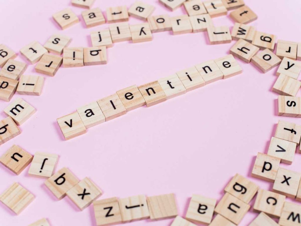 Imagen con letras de Scrabble formando la palabra 'valentine' sobre un fondo rosado, con fichas adicionales dispersas alrededor.