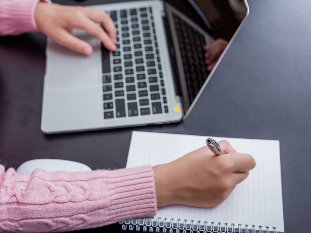 Estudiante escribiendo notas mientras trabaja en su TFG frente a un portátil.