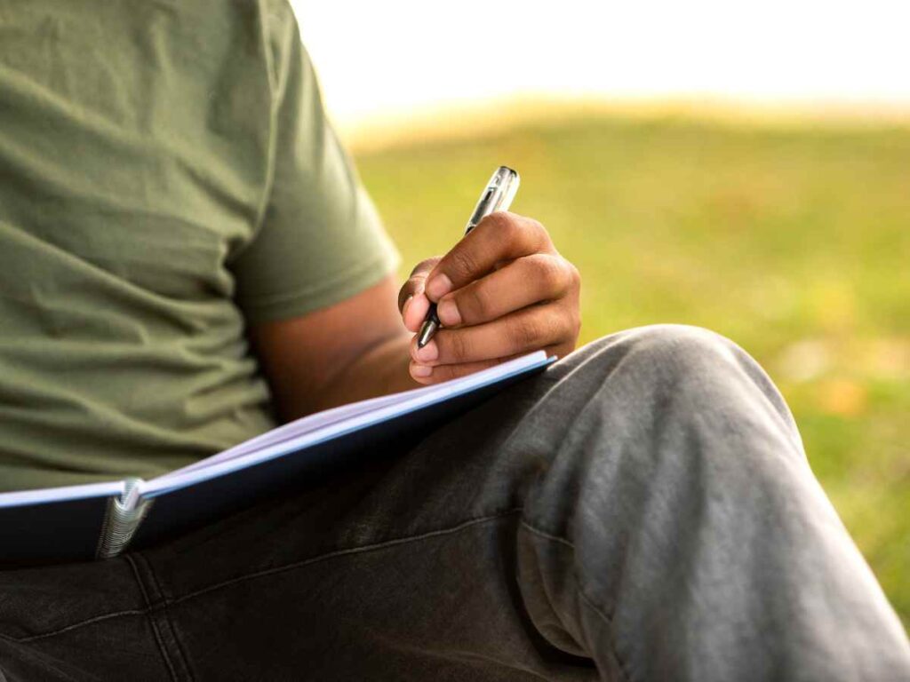 Persona escribiendo en un cuaderno al aire libre, enfocándose en la mano.